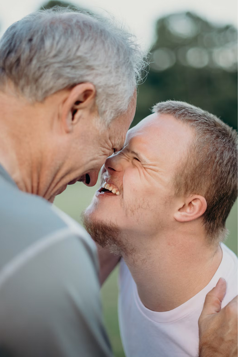 An older man and a younger man laughing together, foreheads touching, outdoors at sunset.