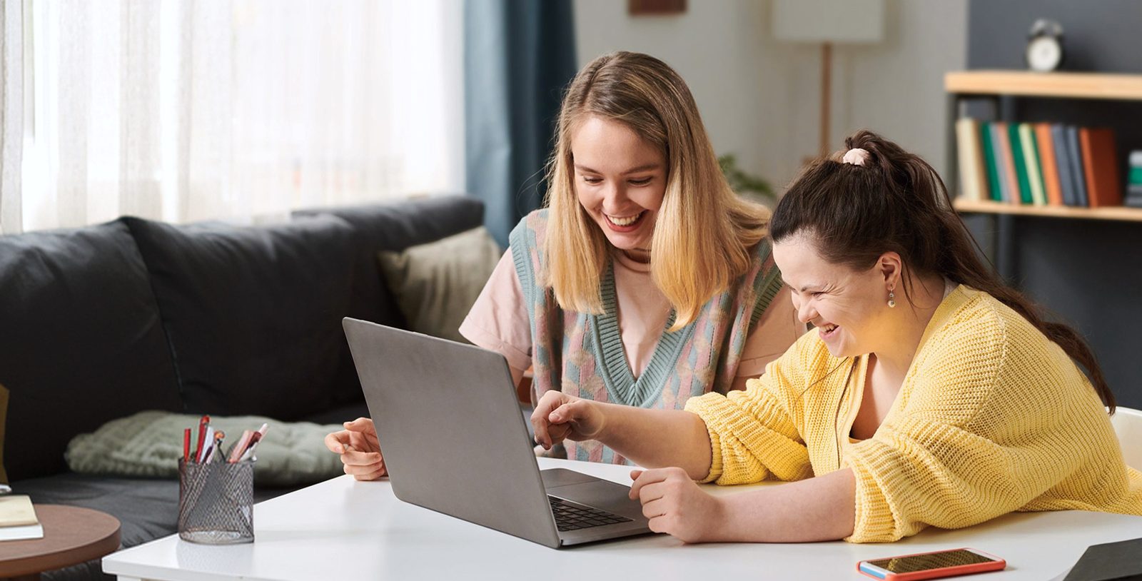 Two young women laughing together at a desk while looking at a laptop in a home setting, with books and stationery nearby.